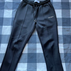 Les deux baller track pants  - Svarta byxor från Les Deux med raka ben och snörning i midjan. Byxorna har diskret logga på ena benet och sydda pressveck framtill för en stilren look. Perfekta för en avslappnad men ändå uppklädd stil.