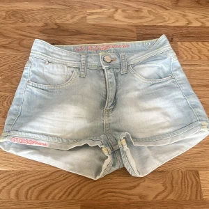 Ljusblå jeansshorts från Guess - Säljer ett par ljusblå jeansshorts från Guess med klassisk femficksdesign och uppvikta benslut. Shortsen har broderad logga i rosa och vit på linningen och bakfickan, samt knapp och dragkedja framtill. ( midjemått 64cm) säljer då de va för små