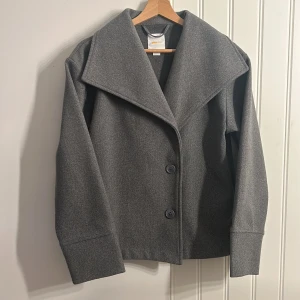 Grå ullkappa med bred krage - Stilren grå kappa med bred krage och två knappar framtill. Jackan har lång ärm och är tillverkad i ett mjukt ullmaterial. Perfekt för kyliga dagar och ger en klassisk look.