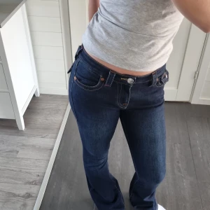 Mörkblå bootcut jeans från True Religion - Säljer ett par mörkblå jeans från True Religion, modell Becca Mid Rise Bootcut. Jeansen har snygga kontrastsömmar, klassiska fickor med lock och knappdetaljer samt en bootcut passform.