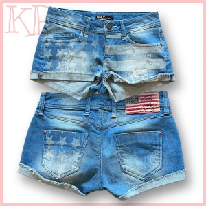 Blå jeansshorts med amerikanskt flaggmönster från ONLY - Säljer ett par blå jeansshorts från ONLY med slitna detaljer och amerikanskt flaggmönster både fram och bak. Shortsen har klassisk femficksdesign, uppvikta benslut och patch med flagga på baksidan. Perfekta för sommaren!