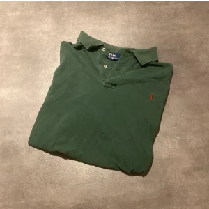 Grön pikétröja från Polo Ralph Lauren - Säljer en klassisk grön pikétröja från Polo Ralph Lauren med röd broderad logga på bröstet. Tröjan har korta ärmar, krage och knappar vid halsen. Perfekt för en stilren och avslappnad look. Tröjan är storlek 2XB och ganska stor i storleken. Bara att höra av sig vid funderingar eller frågor!😀