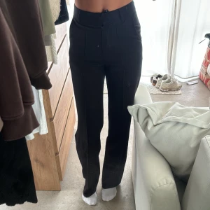 Svarta kostymbyxor med pressveck från madlady  - Snygga svarta kostymbyxor med hög midja och raka ben. Byxorna har pressveck framtill, hällor för bälte och stängs med dragkedja och knapp. Perfekta för en stilren look.