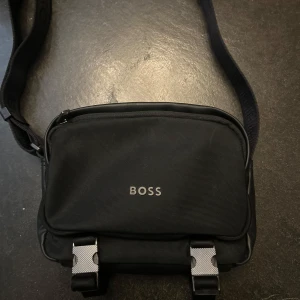 Svart axelväska från BOSS - Snygg svart axelväska från BOSS med justerbar rem och tydlig logga både på väskan och remmen. Väskan har dragkedja upptill och två spännen framtill. 