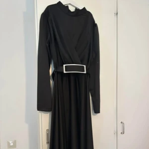Svart långklänning med bälte - Snygg svart långklänning med lång ärm och markerad midja tack vare ett brett bälte med spänne. Klänningen har en enkel och stilren design med rund halsringning och ett mjukt, följsamt tyg.