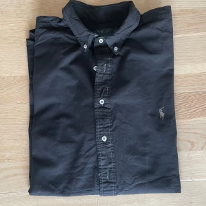 Svart skjorta från Polo Ralph Lauren - Klassisk svart skjorta från Polo Ralph Lauren i bomull. Skjortan har knappar framtill, krage och den ikoniska broderade loggan på bröstet. Perfekt för dig som gillar stilrena och tidlösa plagg med snygg passform.