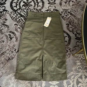 Olivgrön långkjol från Part Two - Snygg olivgrön långkjol från Part Two med klassisk rak passform, bälteshällor och två bakfickor. Kjolen har en slits framtill för extra rörelsefrihet och är tillverkad i mjuk bomull. Perfekt för dig som gillar stilren och enkel design. Helt oanvänd, nypris: 800 kr