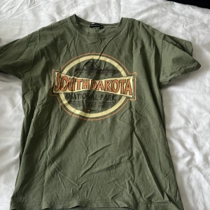 Olivgrön South Dakota t-shirt PLT - Olivgrön t-shirt från PrettyLittleThing med coolt South Dakota National Park-tryck framtill. Klassisk rund halsringning och korta ärmar. Skön och avslappnad passform, perfekt till jeans eller shorts. Tillverkad i mjuk bomull.