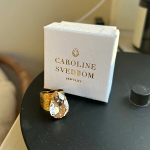 Guldfärgad ring stor kristall Caroline Svedbom - Säljer en statementring från Caroline Svedbom i guldfärgad metall med en stor, droppformad klar kristall. Ringen har en bred, räfflad ringbas som ger en lyxig känsla och kristallen fångar ljuset superfint. Kristallen är i färgen ”Silk” och liknar färgen crystal men med en lite varmare ton är crystal💓