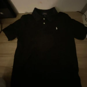 Svart pikétröja från Polo Ralph Lauren - Klassisk svart pikétröja från Polo Ralph Lauren med vit broderad logga på bröstet. Tröjan har korta ärmar, krage och knappar framtill. Perfekt för en stilren och tidlös look. 8,5/10 skick