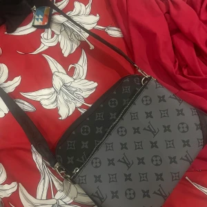 Louis Vuitton Trio Bag - Säljer en snygg grå axelväska från Louis Vuitton med klassiskt monogrammönster. Väskan har svarta detaljer och en bred axelrem. Perfekt för att bära dina viktigaste saker med stil. Eventuella frågor så är det bara att ta kontakt :)
