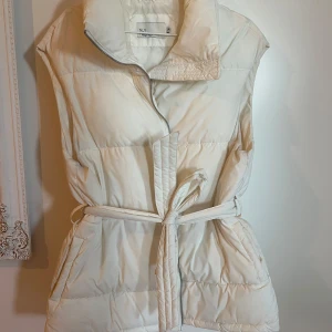Vit dunväst med bälte från NLY Trend - Säljer en vit dunväst från NLY Trend med hög krage, dragkedja och knytskärp i midjan. Västen har en stilren look och är perfekt för lager-på-lager. Passar dig som gillar en modern och enkel stil. Har en liten fläck som syns på sista bilden. Går säkert att få bort om man orkar. 
