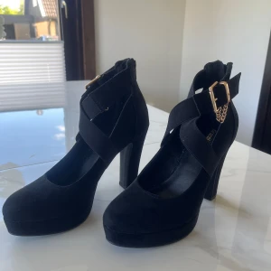 Svarta pumps med platå och bred klack från Graceland - Säljer ett par svarta pumps med bred klack. Skorna har korsade remmar över foten och ett dekorativt guldfärgad spänne vid ankeln. De är använda 1 gång,är då egentligen som ny och i storlek 38.