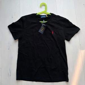 Svart t-shirt från Polo Ralph Lauren - Svart t-shirt från Polo Ralph Lauren med klassisk rund hals och röd broderad logga på bröstet. Enkel och stilren design, perfekt för en avslappnad look. Perfekt nu till sommaren!