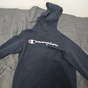 Mörkblå hoodie från Champion - Säljer en mörkblå hoodie från Champion med stor logga på bröstet. Tröjan har huva och en stor magficka framtill. Perfekt för en avslappnad stil och enkel att matcha med jeans eller träningsbyxor.