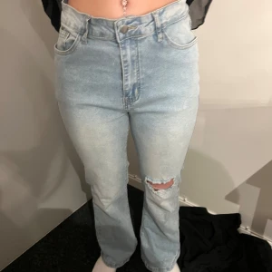 Ljusblå bootcut jeans med slitning - Säljer ett par ljusblå jeans med bootcut-modell och hög midja. Jeansen har en snygg slitning på ena knät och klassiska fem fickor. Perfekta för en avslappnad och trendig look.