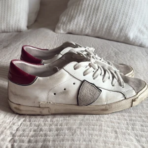 Philippe Model vita sneakers med vinröd detalj - Snygga vita sneakers från Philippe Model med vinröda detaljer på hälen och grå mocka på tån. Skorna har snörning, rund tå och en platt sula. Tillverkade i skinn och mocka för en lyxig känsla och cool look. Man får också med boxen!