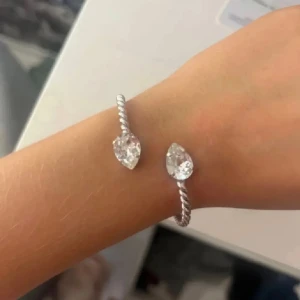 Caroline swedbom  - Snyggt armband i silverfärg med tvinnad design och två stora, droppformade glittriga stenar i varje ände. Perfekt statement piece som drar blickarna till sig och ger en lyxig känsla till din outfit. 