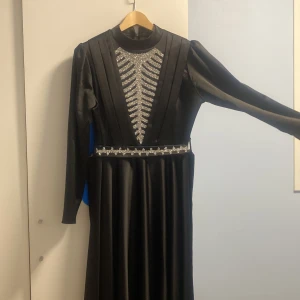 Svart långklänning med glitterdetaljer - Snygg svart långklänning från Aycan med glittrande silverdetaljer framtill och i midjan. Klänningen har lång ärm, hög hals och är tillverkad i ett glansigt material som ger en lyxig känsla. Perfekt för dig som vill sticka ut med elegans.