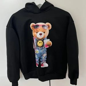 Svart hoodie med nalletryck från SHEIN - Svart hoodie från SHEIN med en stor färgglad nalle på framsidan. Nallen har rosa solglasögon, smileytröja, jeans och sneakers. Hoodien har huva och långa ärmar, perfekt för dig som gillar coola och lekfulla prints.