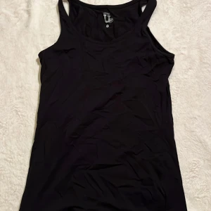 Svart linne - Svart, klassiskt linne från Basic U i mjuk bomull. Modellen är ärmlös med breda axelband och rund halsringning. Perfekt att styla till jeans eller shorts för en enkel och clean look.