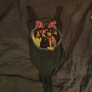 Svart AC/DC bodysuit med tryck - Svart bodysuit med AC/DC-tryck framtill och coolt bandmotiv i färg. Djup v-ringning med tre svarta band över bröstet för extra edge. Ärmlös modell med breda axelband och figurnära passform. Perfekt för dig som vill sticka ut med rockig stil.