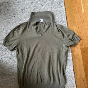 Olivgrön pikétröja från Bläck - Snygg olivgrön pikétröja från Bläck med klassisk krage och knappar framtill. Tröjan har korta ärmar och är tillverkad i mjuk bomull, perfekt för en avslappnad och stilren look. Passar dig som gillar enkel och clean stil.