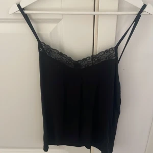 Svart linnetopp med spetskant - Snygg svart linnetopp med tunna axelband och dekorativ spets längs urringningen. Perfekt att styla lager på lager eller bära som den är. Enkel och stilren design som passar till många olika looks.