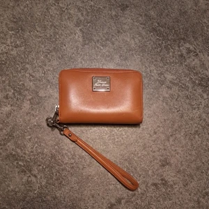 Brun plånbok/väska - Snygg brun plånbok med silverfärgad logoplatta framtill. Plånboken har avtagbar handledsrem och dragkedja runt om. Insidan har flera kortfack och plats för mynt och sedlar. Perfekt att bära i handen eller slänga ner i väskan. Använd men mycket bra skick. Bundlerabatt finns 😊