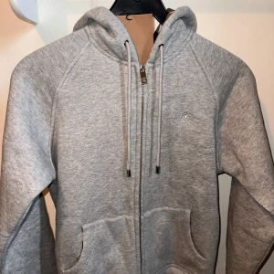 Grå hoodie med dragkedja från Burberry - Grå hoodie från Burberry med dragkedja framtill, huva med snörning och broderad logga på bröstet. Mjuk bomullskänsla och känguruficka framtill. Perfekt för en chill och stilren look.