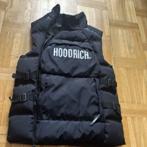 Svart dunväst från Hoodrich XS - Svart dunväst från Hoodrich i storlek XS. Västen har en hög krage, justerbara spännen på sidorna och stor vit logga på bröstet. Yttermaterialet är glansigt och insidan har mjukt foder vid nacken. Perfekt för lager-på-lager och streetwear.