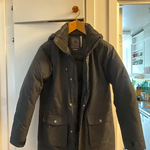 Svart dunjacka från Everest med huva - Svart dunjacka från Everest med huva, dragkedja och tryckknappar framtill. Jackan har stora fickor med lock och en bröstficka med dragkedja. Hittade en liten röd fläck vid ena fickan, men skulle tro att den går bort i tvätten. Den är i barnstorlek men passar även en S. 