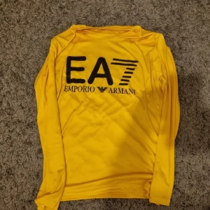 Gul långärmad tröja från Emporio Armani EA7 - Säljer en gul långärmad tröja från Emporio Armani EA7 med stor svart logga på bröstet. Tröjan har en sportig look och är tillverkad i ett mjukt, stretchigt material som passar perfekt till träning eller vardag.