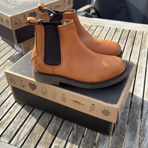 Bruna chelsea boots i skinn sneaky steve - Säljer ett par snygga bruna chelsea boots från sneaky steve i skinn med elastiska svarta paneler på sidorna och fordring inuti. Perfekta för dig som vill ha en klassisk och stilren look till hösten eller våren.