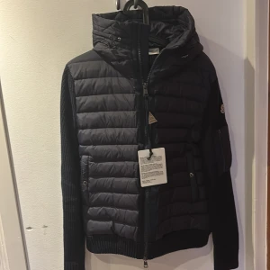Svart pufferjacka från Moncler - Säljer en svart pufferjacka från Moncler med stickade ärmar och huva. Jackan har dragkedja framtill, ribbade muddar och Moncler-logga på ärmen. Perfekt för kyliga dagar och riktigt snygg streetstil.