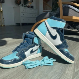 Nike Air Jordan 1 Retro High OG blå/vit - Säljer ett par Nike Air Jordan 1 Retro High OG i blått och vitt. Skorna har högt skaft, klassisk Nike swoosh och perforerad tå. Färgerna är ljusblå, marinblå och vit. Medföljer extra blå skosnören. Perfekta för dig som gillar streetwear och sneakers.