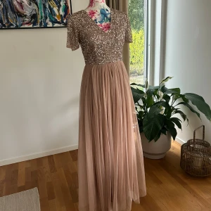 Champagnefärgad långklänning med paljetter - Säljer en elegant champagnefärgad långklänning med glittrig paljettöverdel och v-ringning. Klänningen har korta ärmar och ett mjukt, flowy kjolparti i tunt tyg. Perfekt för speciella tillfällen!