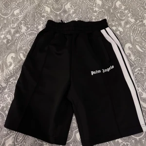 Svarta shorts från Palm Angels med vita ränder - Svarta shorts från Palm Angels med vita sidoremsor och logga tryckt på vänster ben. Shortsen har elastisk midja och sidofickor. Perfekta för en sportig och trendig look.