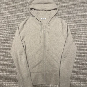 Grå hoodie i kashmir från Soft Goat - Mjuk och lyxig grå hoodie från Soft Goat i kashmir. Tröjan har dragkedja framtill, huva med dragsko och ribbade muddar. Perfekt för en avslappnad och stilren look.