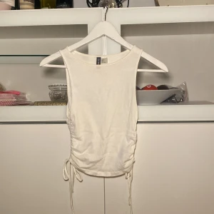 Vit ribbad linnetopp med knyt - Säljer en vit ribbad linnetopp med knyt i sidorna. Toppen är ärmlös och har en rund halsringning. Perfekt för varma dagar och enkel att matcha med olika outfits.