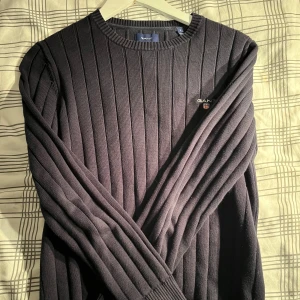 Svart ribbad tröja från GANT - Säljer en svart ribbstickad tröja från GANT i premium bomull. Tröjan har rund halsringning, diskret GANT-logga på bröstet och långa ärmar. Perfekt för dig som gillar klassisk och stilren look med en modern touch.