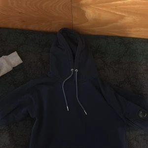 Marinblå hoodie från C.P. Company - Säljer en marinblå hoodie från C.P. Company med klassisk huva och snörning. Tröjan har lång ärm och den ikoniska linsen på vänster ärm. Perfekt för dig som gillar streetwear och vill ha en stilren look.