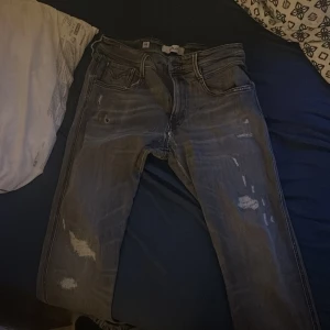 Replay grå jeans med slitningar - Säljer ett par grå jeans från Replay i modell W32 L34. Jeansen har snygga slitningar och en tvättad look, klassiska fem fickor och coola detaljer på bakfickorna. Perfekta för dig som gillar en avslappnad och trendig stil.