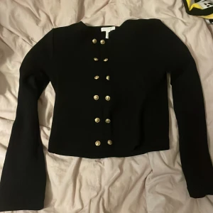 Svart kofta med guldknappar H&M - Stilren svart kofta från H&M med raka axlar och dekorativa dubbla rader av guldknappar framtill. Långärmad modell med något croppad passform, perfekt till jeans eller kjol. Materialet känns mjukt och har en lätt stretch.