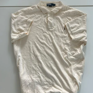 Beige piké från Ralph Lauren  - Klassisk beige pikétröja från Ralph Lauren i storlek XL. Tröjan har korta ärmar, krage och knäppning framtill samt den ikoniska broderade loggan på bröstet. Tillverkad i mjuk bomull som är skön mot huden. Perfekt för en clean och avslappnad stil.