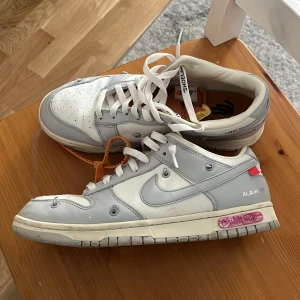 Nike Dunk Low Off-White vit/grå - Nike Dunk Low Off-White i vitt och ljusgrått med klassiska Off-White-detaljer som orange zip-tie och text på sidan. Skorna har vit bas, ljusgrå overlays och rosa accent på sulan. Tjock platt sula, snörning och extra snördetaljer. Perfekt för dig som gillar streetwear.