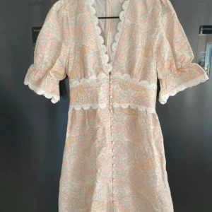 Pastellfärgad kortklänning med spets - Supersöt kortklänning i pastelliga toner med paisleymönster, v-ringning och dekorativa knappar framtill. Klänningen har korta puffärmar och fina spetsdetaljer längs ärmar, midja och halsringning. Perfekt för sommarens alla tillfällen!