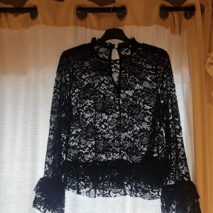 Svart spetsblus med volangdetaljer - Säljer en svart blus i genombruten spets med lång ärm och volangdetaljer vid ärmslut och nederkant. Blusen har hög hals med liten volang och en snygg öppning bak i nacken. Perfekt för dig som gillar romantisk och edgy stil. Kontakta innan köp och fråga om ni har några frågor eller vill ha fler bilder. 
