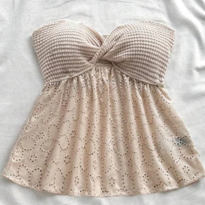 Beige tubtopp med broderade blommor - Söt beige tubtopp med virkad överdel och broderade blommor på den utsvängda nederdelen. Toppen är axelbandslös och har en twistad detalj framtill. Perfekt för varma dagar.