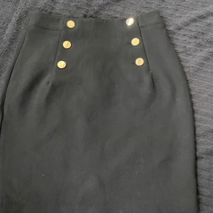 Svart pennkjol med guldknappar - Säljer en svart pennkjol med sex dekorativa guldknappar framtill. Kjolen har hög midja och klassisk, stilren design. Perfekt för dig som gillar en tidlös look med en liten twist.